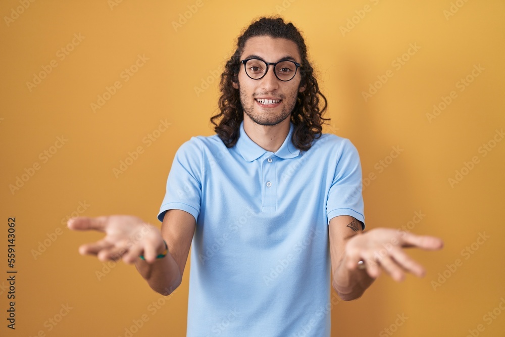 Young hispanic man standing over yellow background smiling cheerful offering hands giving assistance and acceptance. Young hispanic man standing over yellow background smiling cheerful offering hands giving assistance and acceptance.