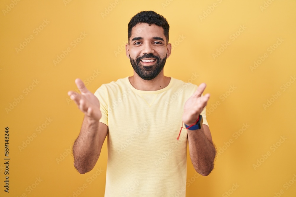 Hispanic man with beard standing over yellow background smiling cheerful offering hands giving assistance and acceptance. Hispanic man with beard standing over yellow background smiling cheerful offering hands giving assistance and acceptance.