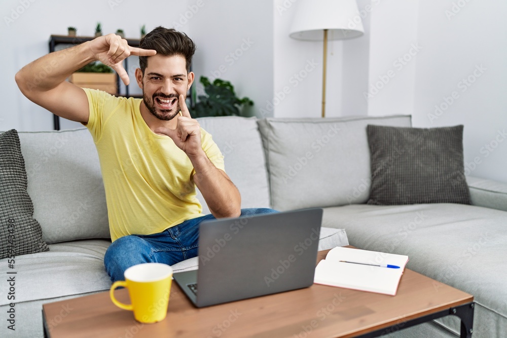 Young man with beard using laptop at home smiling making frame with hands and fingers with happy face. creativity and photography concept. Young man with beard using laptop at home smiling making frame with hands and fingers with happy face. creativity and photography concept.