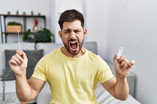 Canvastavla Young man with beard holding coronavirus infection nasal test angry and mad screaming frustrated and furious, shouting with anger