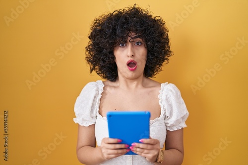 Young brunette woman with curly hair using touchpad over yellow background in shock face, looking skeptical and sarcastic, surprised with open mouth
