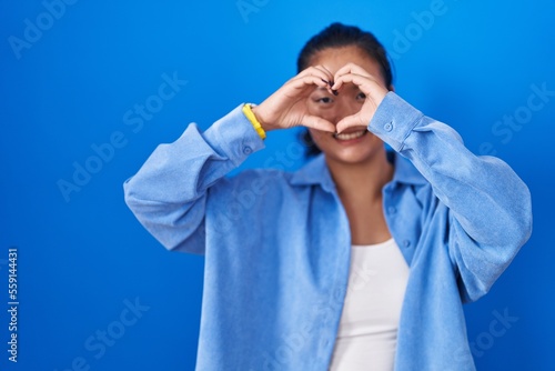 Asian young woman standing over blue background doing heart shape with hand and fingers smiling looking through sign