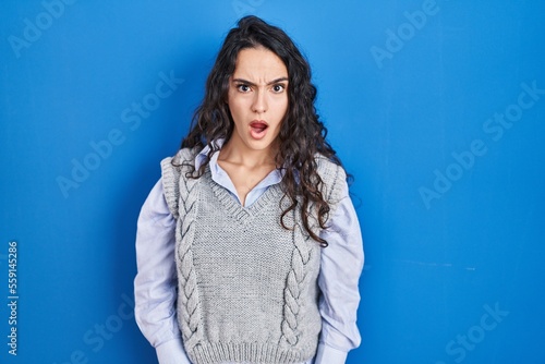 Young brunette woman standing over blue background in shock face, looking skeptical and sarcastic, surprised with open mouth