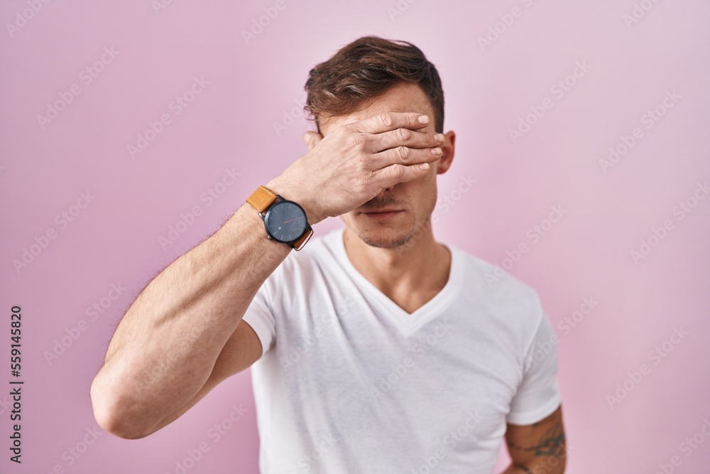 Fototapeta premium Caucasian man standing over pink background covering eyes with hand, looking serious and sad. sightless, hiding and rejection concept