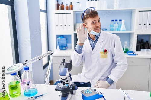 Caucasian man working at scientist laboratory smiling with hand over ear listening and hearing to rumor or gossip. deafness concept.