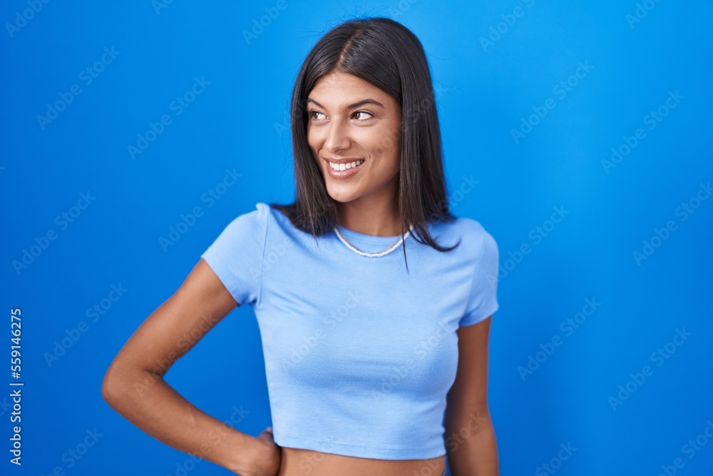 Brunette young woman standing over blue background looking away to side with smile on face, natural expression. laughing confident. Brunette young woman standing over blue background looking away to side with smile on face, natural expression. laughing confident.