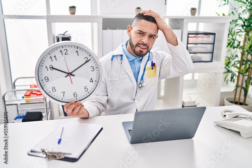 Hispanic doctor man holding clock at the clinic stressed and frustrated with hand on head, surprised and angry face