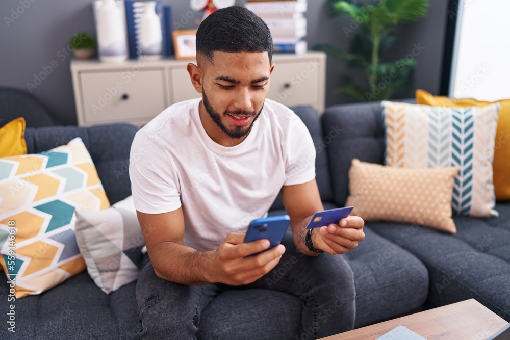 Young latin man using smartphone and credit card sitting on sofa at home Young latin man using smartphone and credit card sitting on sofa at home