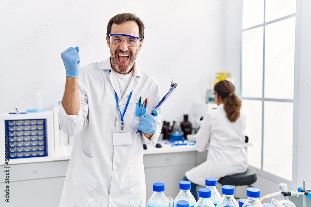 Middle age man working at scientist laboratory screaming proud, celebrating victory and success very excited with raised arms