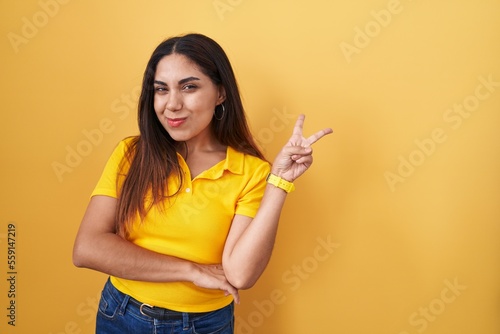 Young arab woman standing over yellow background smiling with happy face winking at the camera doing victory sign with fingers. number two.