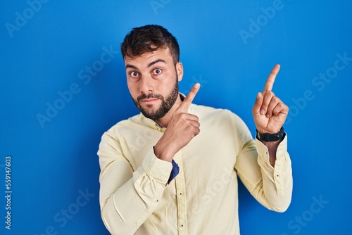 Handsome hispanic man standing over blue background pointing aside worried and nervous with both hands, concerned and surprised expression