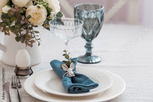 Beautiful spring table setting. A plate with a cotton napkin with a bunny. Silverware and a vase of flowers on a linen tablecloth. The concept of festive serving and a bright Easter holiday.