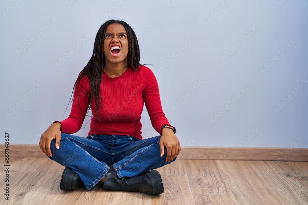 Young african american with braids sitting on the floor at home angry and mad screaming frustrated and furious, shouting with anger. rage and aggressive concept.