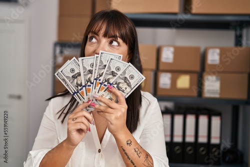 Young beautiful hispanic woman ecommerce business worker holding dollars over mouth at office