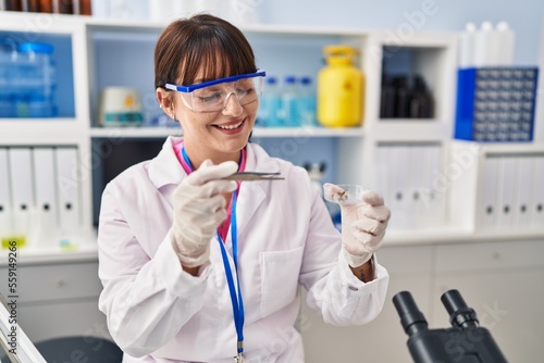 Young beautiful hispanic woman scientist smiling confident holding plant sample and tweezers at pharmacy