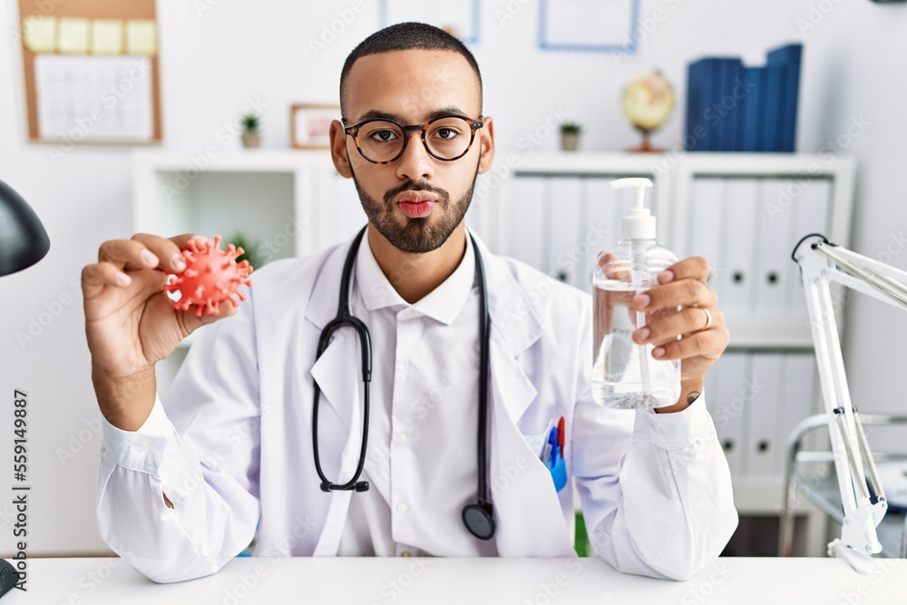 African american doctor man holding virus toy an sanitizer gel looking at the camera blowing a kiss being lovely and sexy. love expression. African american doctor man holding virus toy an sanitizer gel looking at the camera blowing a kiss being lovely and sexy. love expression.
