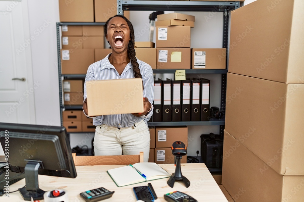 African woman working at small business ecommerce holding box angry and mad screaming frustrated and furious, shouting with anger looking up.