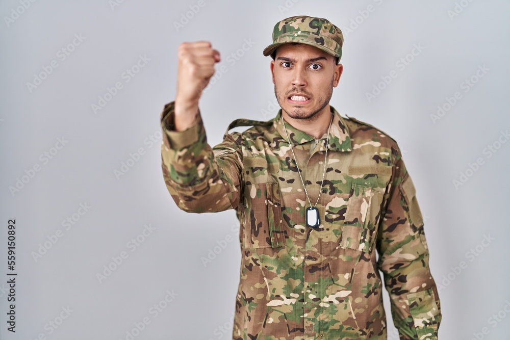 Young hispanic man wearing camouflage army uniform angry and mad raising fist frustrated and furious while shouting with anger. rage and aggressive concept. Young hispanic man wearing camouflage army uniform angry and mad raising fist frustrated and furious while shouting with anger. rage and aggressive concept.