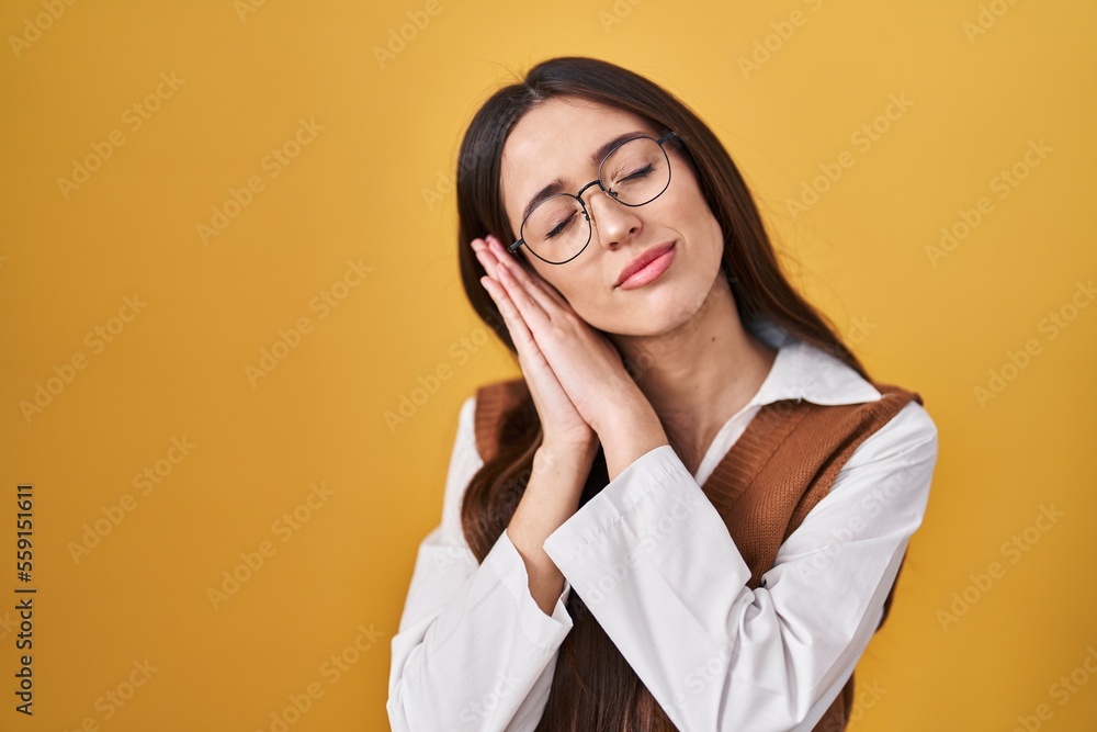 Young brunette woman standing over yellow background wearing glasses sleeping tired dreaming and posing with hands together while smiling with closed eyes.