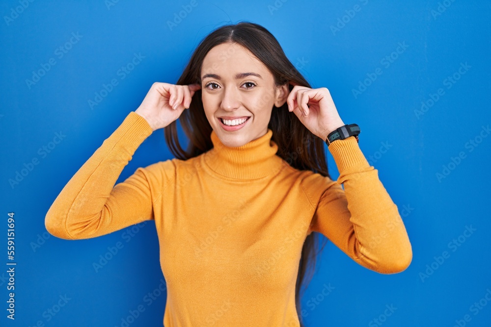 Young brunette woman standing over blue background smiling pulling ears with fingers, funny gesture. audition problem Young brunette woman standing over blue background smiling pulling ears with fingers, funny gesture. audition problem