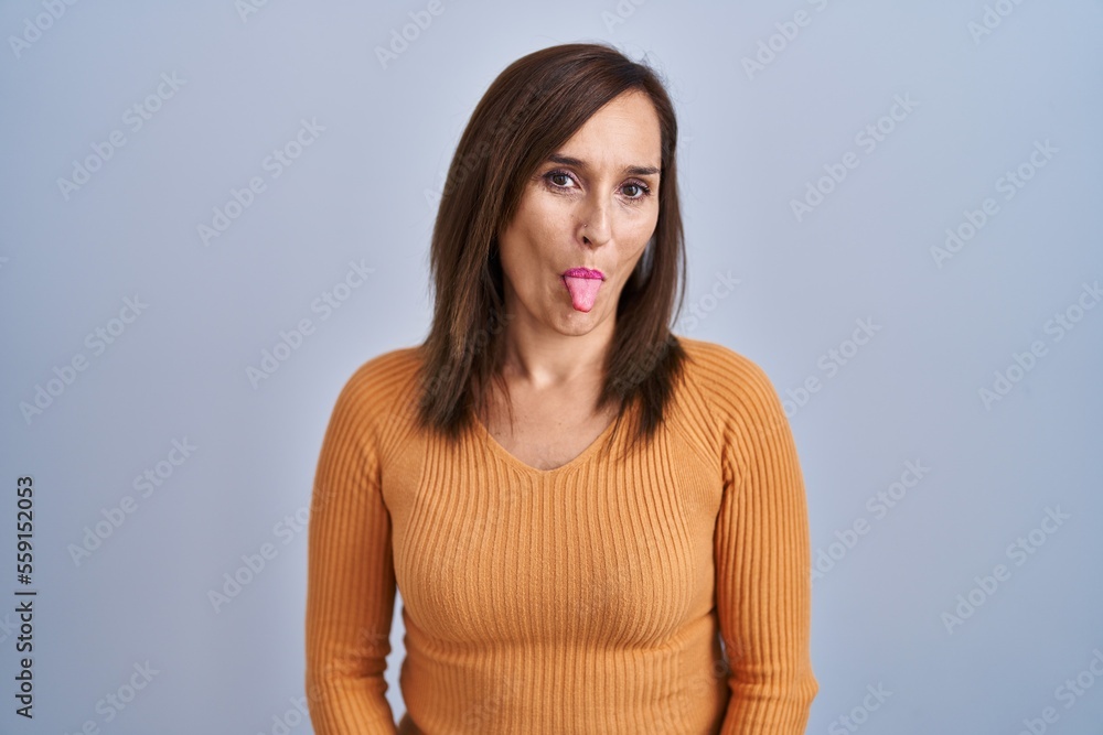 Middle age brunette woman standing wearing orange sweater sticking tongue out happy with funny expression. emotion concept. Middle age brunette woman standing wearing orange sweater sticking tongue out happy with funny expression. emotion concept.