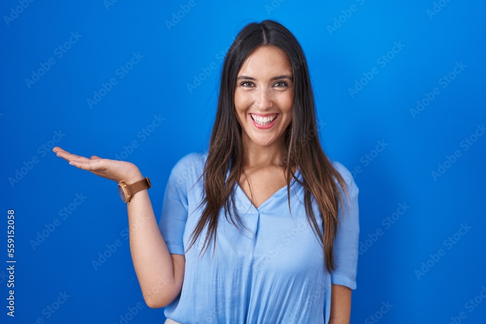 Young brunette woman standing over blue background smiling cheerful presenting and pointing with palm of hand looking at the camera. Young brunette woman standing over blue background smiling cheerful presenting and pointing with palm of hand looking at the camera.