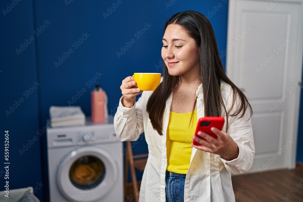 Young hispanic woman using smartphone and drinking coffee waiting for washing machine at laundry room Young hispanic woman using smartphone and drinking coffee waiting for washing machine at laundry room