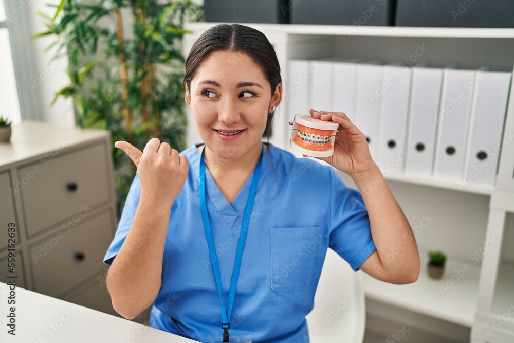 Young latin dentist woman holding denture with braces pointing thumb up to the side smiling happy with open mouth