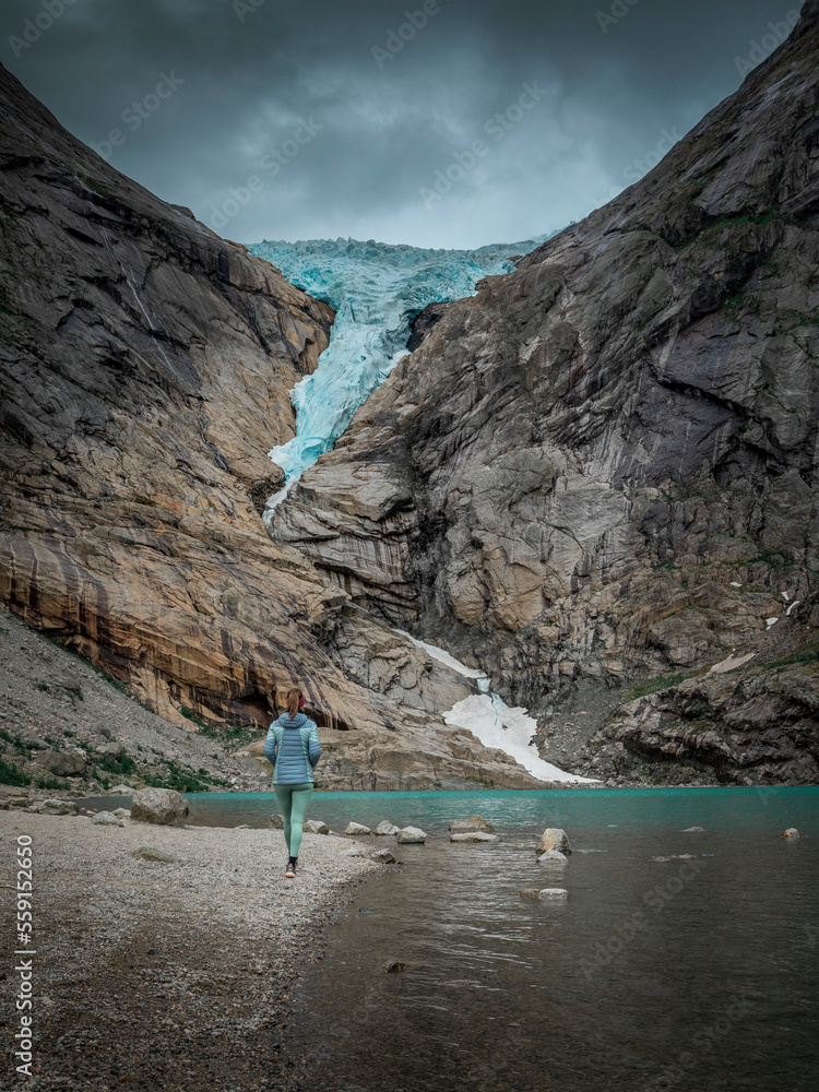 Woman hiking along beach of glacier lake at Briksdalsbreen glacier in the mountains of Jostedalsbreen national park in Norway, turquoise water and blue ice