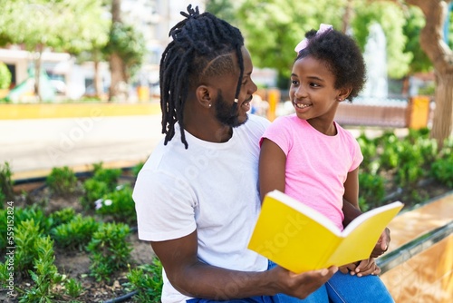 Father and daughter reading book sitting together on bench at park