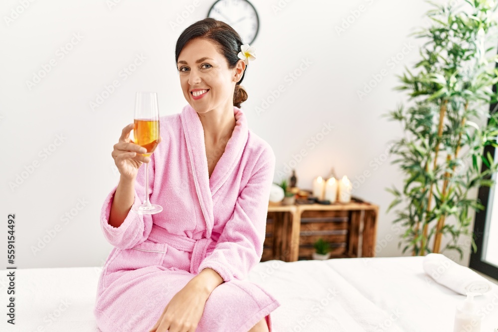 Middle age hispanic woman toasting with champagne sitting on massage table at beauty center Middle age hispanic woman toasting with champagne sitting on massage table at beauty center