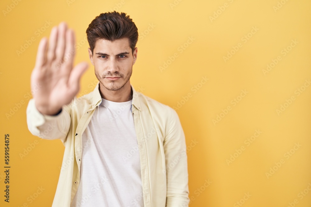 Young hispanic man standing over yellow background doing stop sing with palm of the hand. warning expression with negative and serious gesture on the face. Young hispanic man standing over yellow background doing stop sing with palm of the hand. warning expression with negative and serious gesture on the face.