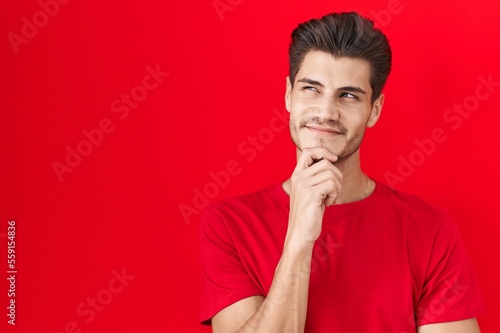 Young hispanic man standing over red background with hand on chin thinking about question, pensive expression. smiling and thoughtful face. doubt concept. © Krakenimages.com