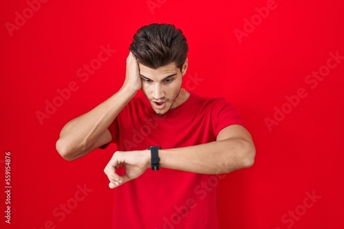 Young hispanic man standing over red background looking at the watch time worried, afraid of getting late