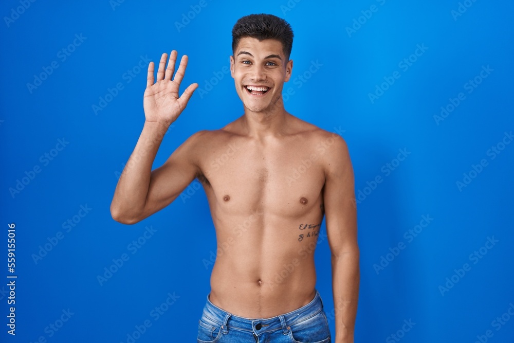 Young hispanic man standing shirtless over blue background waiving saying hello happy and smiling, friendly welcome gesture