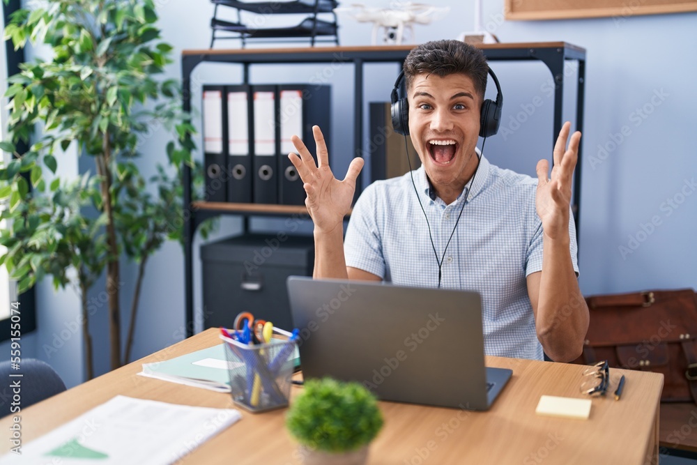 Young hispanic man working at the office wearing headphones celebrating crazy and amazed for success with arms raised and open eyes screaming excited. winner concept Young hispanic man working at the office wearing headphones celebrating crazy and amazed for success with arms raised and open eyes screaming excited. winner concept