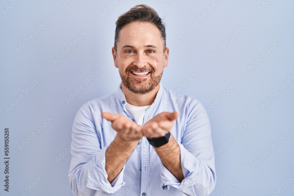Middle age caucasian man standing over blue background smiling with hands palms together receiving or giving gesture. hold and protection Middle age caucasian man standing over blue background smiling with hands palms together receiving or giving gesture. hold and protection