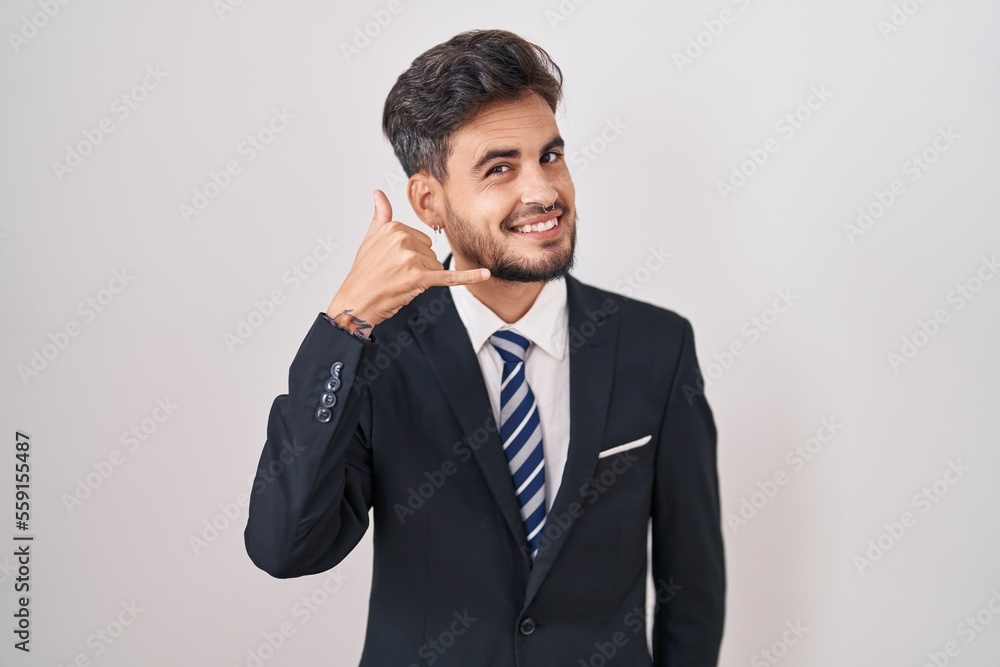 Young hispanic man with tattoos wearing business suit and tie smiling doing phone gesture with hand and fingers like talking on the telephone. communicating concepts. Young hispanic man with tattoos wearing business suit and tie smiling doing phone gesture with hand and fingers like talking on the telephone. communicating concepts.