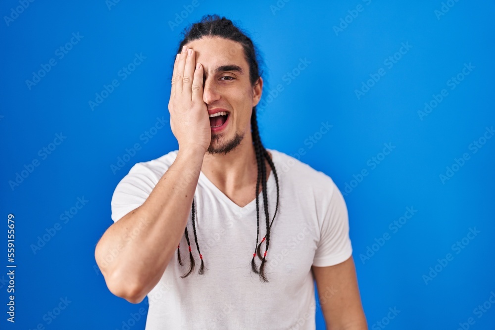 Hispanic man with long hair standing over blue background covering one eye with hand, confident smile on face and surprise emotion. Hispanic man with long hair standing over blue background covering one eye with hand, confident smile on face and surprise emotion.