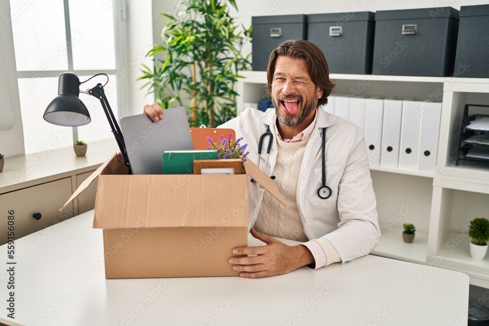 Handsome middle age doctor man holding box with items sticking tongue out happy with funny expression. - obrazy, fototapety, plakaty 