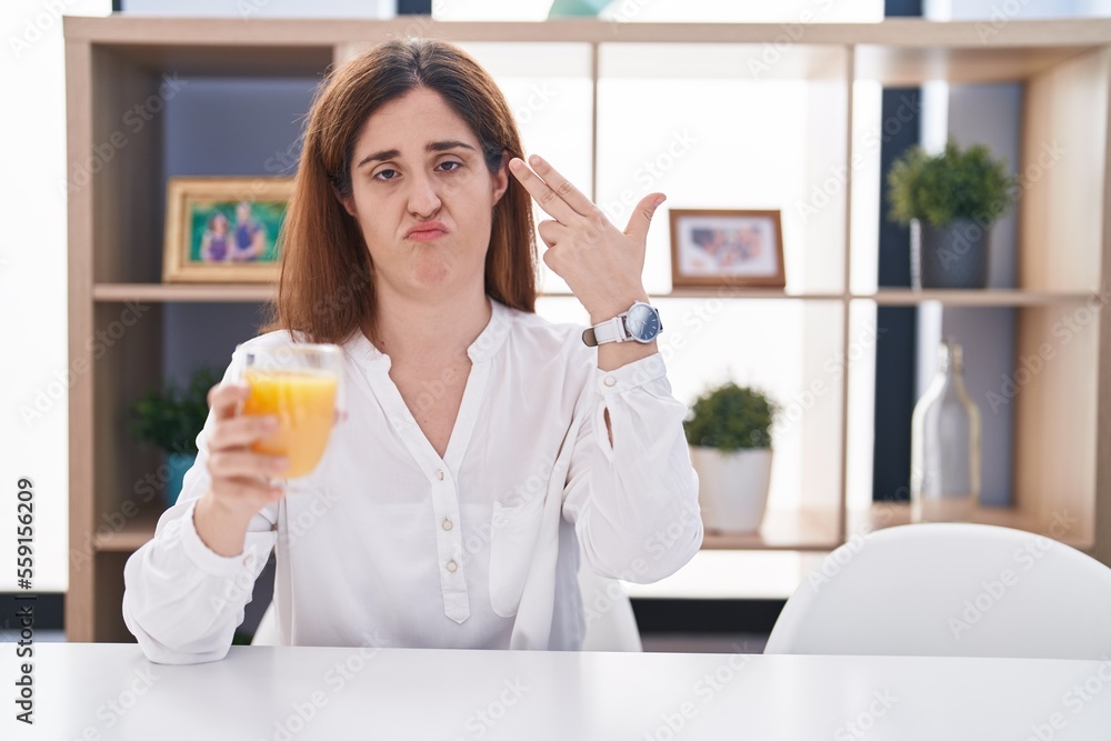 Brunette woman drinking glass of orange juice shooting and killing oneself pointing hand and fingers to head like gun, suicide gesture. Brunette woman drinking glass of orange juice shooting and killing oneself pointing hand and fingers to head like gun, suicide gesture.