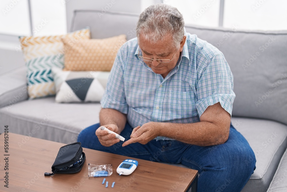 Middle age grey-haired man measuring glucose sitting on sofa at home Middle age grey-haired man measuring glucose sitting on sofa at home