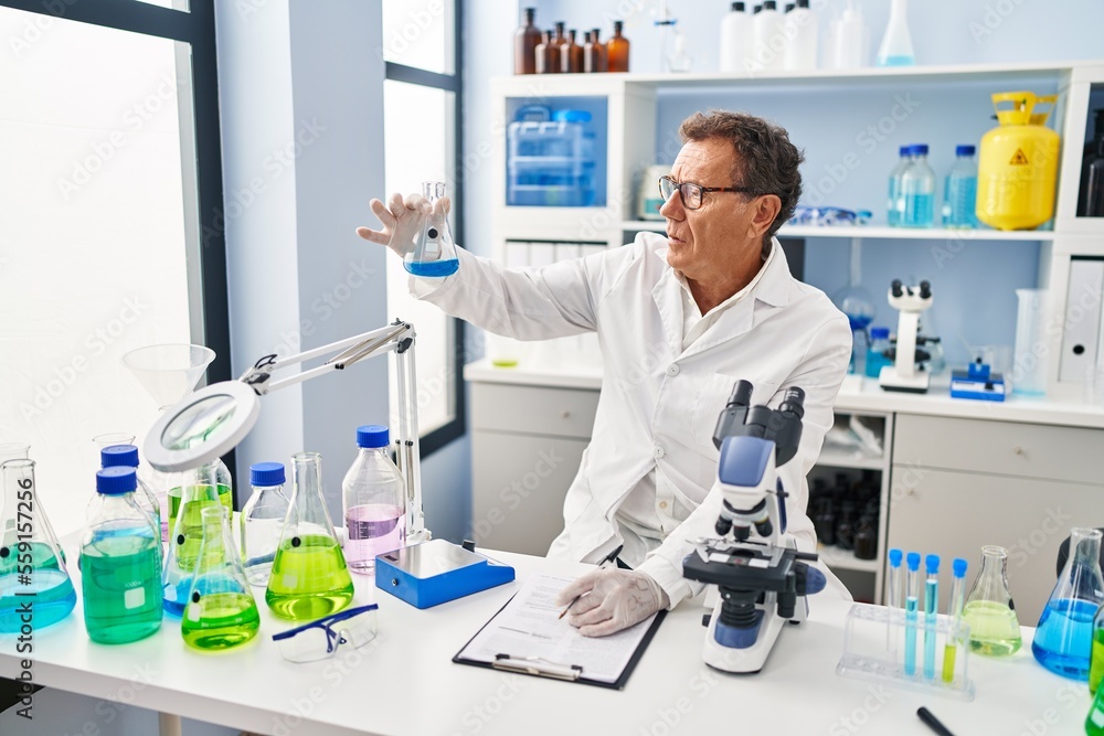 Middle age man wearing scientist uniform looking test tube writing on clipboard at laboratory Middle age man wearing scientist uniform looking test tube writing on clipboard at laboratory