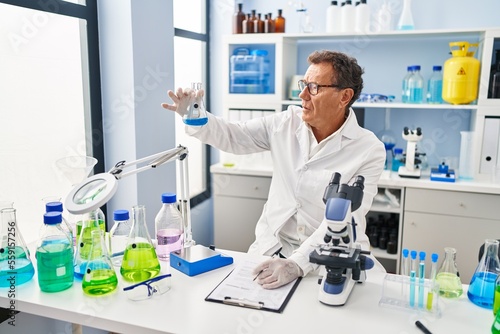 Middle age man wearing scientist uniform looking test tube writing on clipboard at laboratory