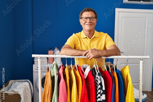 Middle age man smiling confident leaning on clothes rack at laundry room