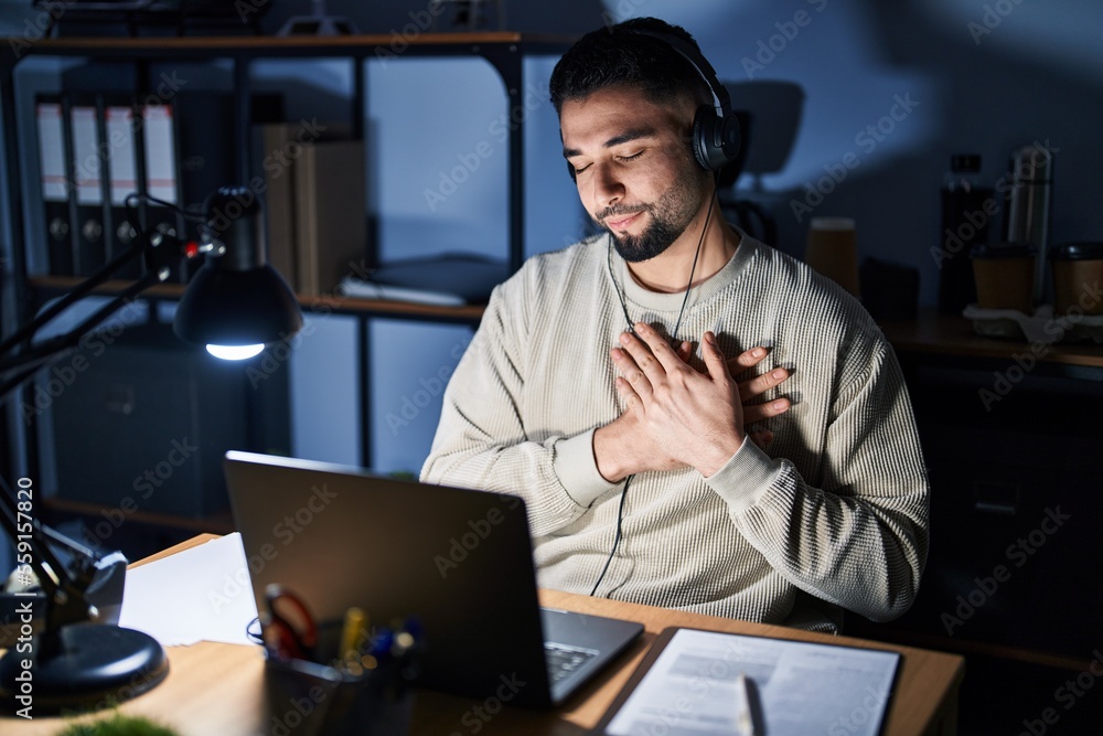 Young handsome man working using computer laptop at night smiling with hands on chest with closed eyes and grateful gesture on face. health concept. Young handsome man working using computer laptop at night smiling with hands on chest with closed eyes and grateful gesture on face. health concept.