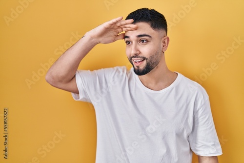 Young handsome man wearing casual t shirt over yellow background very happy and smiling looking far away with hand over head. searching concept.