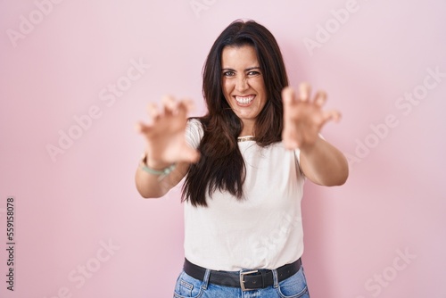 Young brunette woman standing over pink background smiling funny doing claw gesture as cat, aggressive and sexy expression