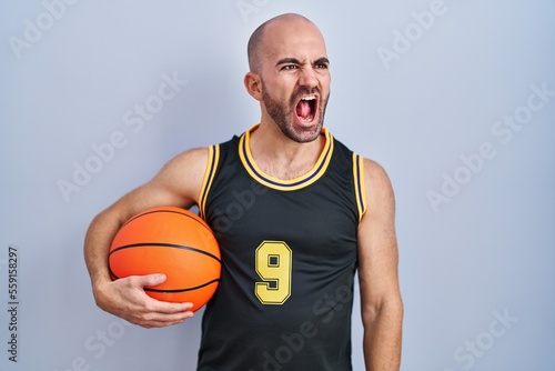 Young bald man with beard wearing basketball uniform holding ball angry and mad screaming frustrated and furious, shouting with anger. rage and aggressive concept.