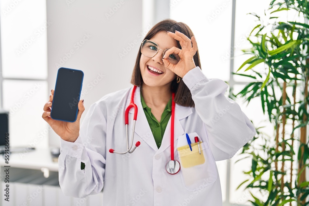 Young hispanic doctor woman showing smartphone screen smiling happy doing ok sign with hand on eye looking through fingers Young hispanic doctor woman showing smartphone screen smiling happy doing ok sign with hand on eye looking through fingers